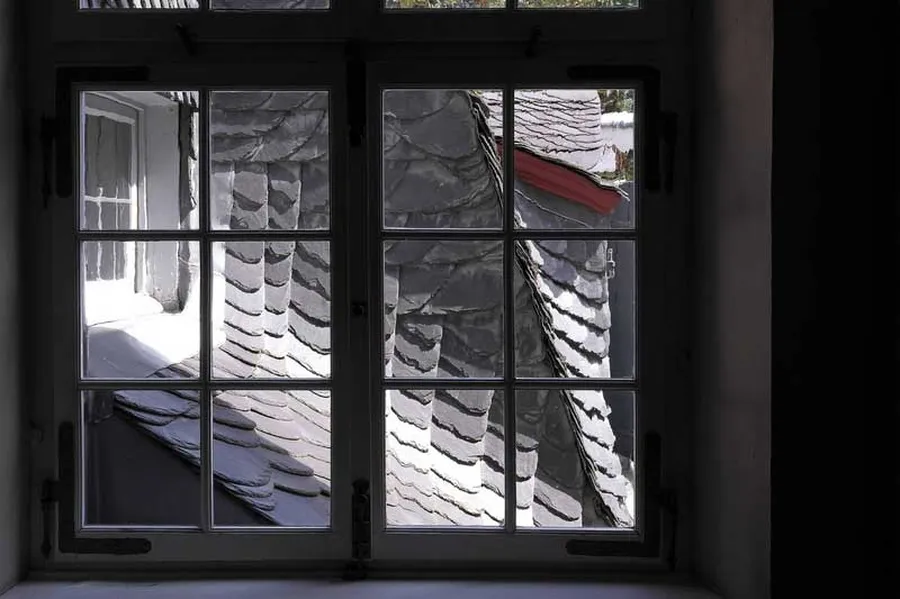 View through a mullioned window of the neighbouring house onto an attic and the sunlit slate roof of the museum building.
