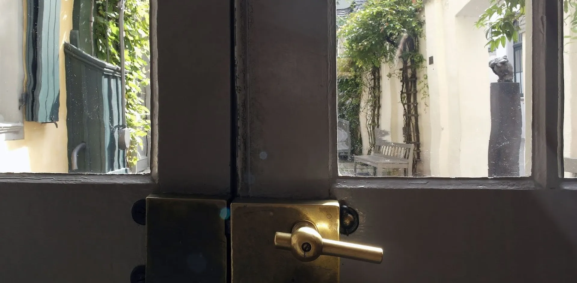 A ray of light falls on the brass fitting of a door to the museum courtyard. Through the two door windows you can see part of the façade of Beethoven's birthplace on the left and a wooden bench in front of a wall with climbing plants on the right.