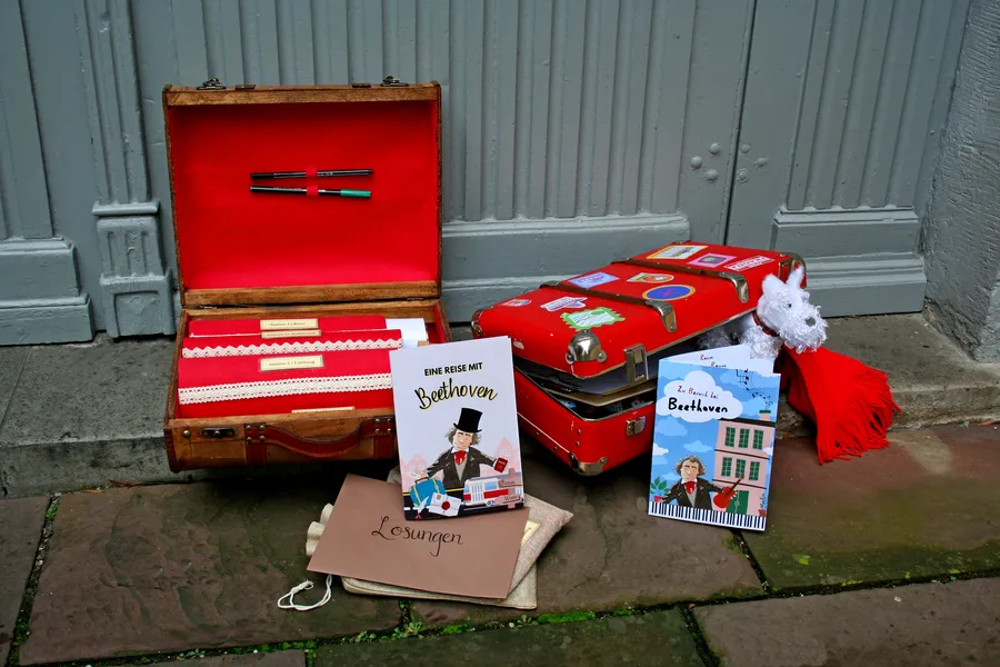 Two red suitcases, one is open, with utensils, including a white dog, a red scarf, 2 Beethoven books.